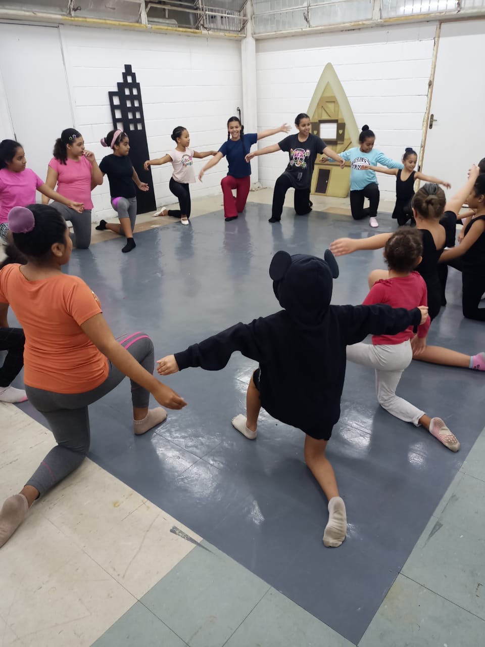 Crianças e jovens em roda, ajoelhadas com os braços abertos, durante aula de balé em sala com piso de linóleo