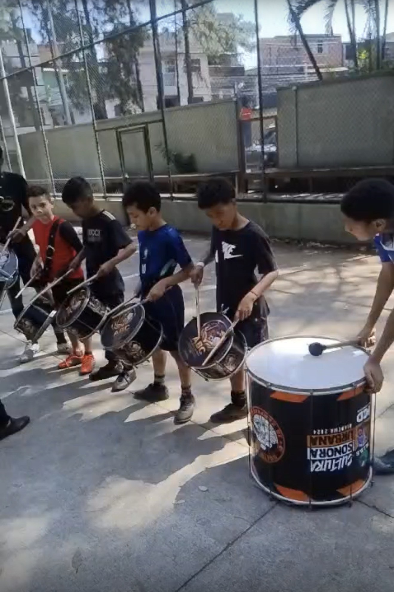 Crianças em fila tocando caixas de percussão em uma quadra ao ar livre, com um surdo do projeto Cultura Sonora Urbana em primeiro plano