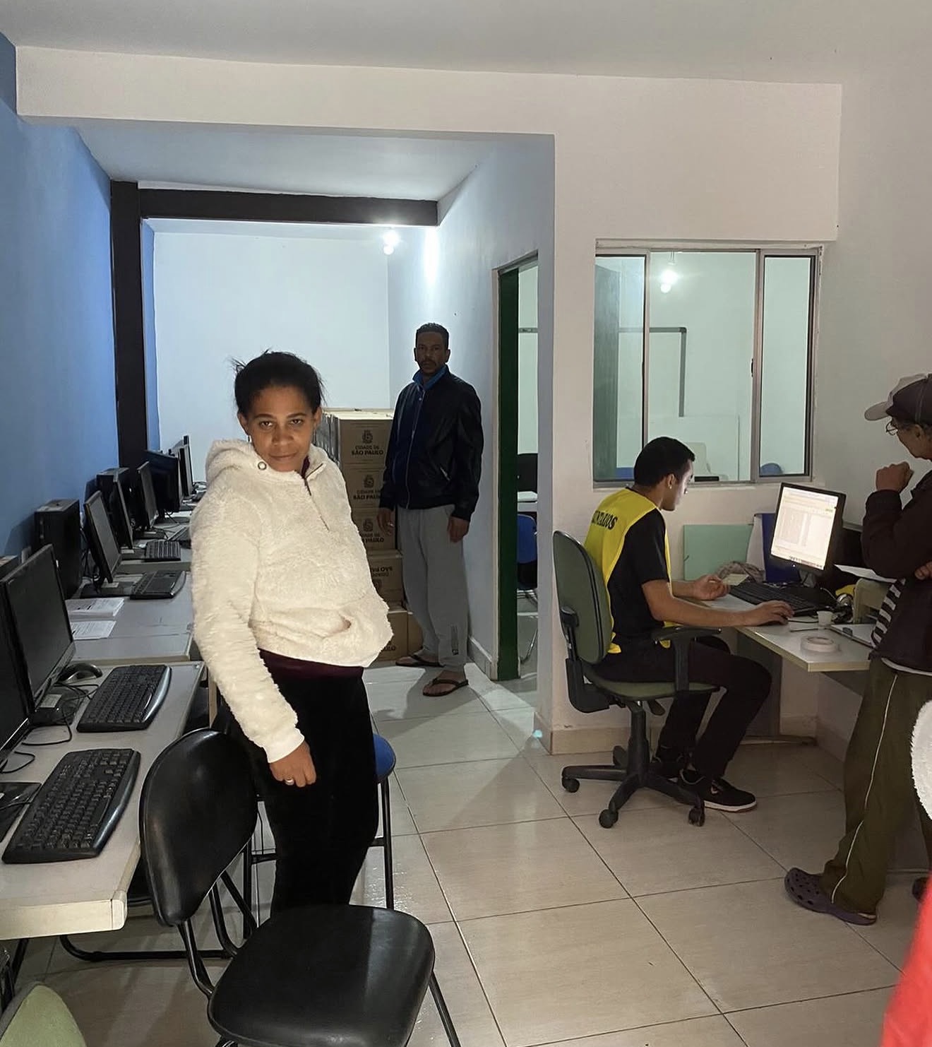 Sala com computadores enfileirados, participantes no ambiente e atendente usando um computador